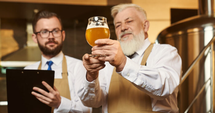 Two professional men wearing white shirts and aprons keeping glass of light beer and examining quality of alcohol. Colleagues looking at drink and writing in folder. Concept of brewery.